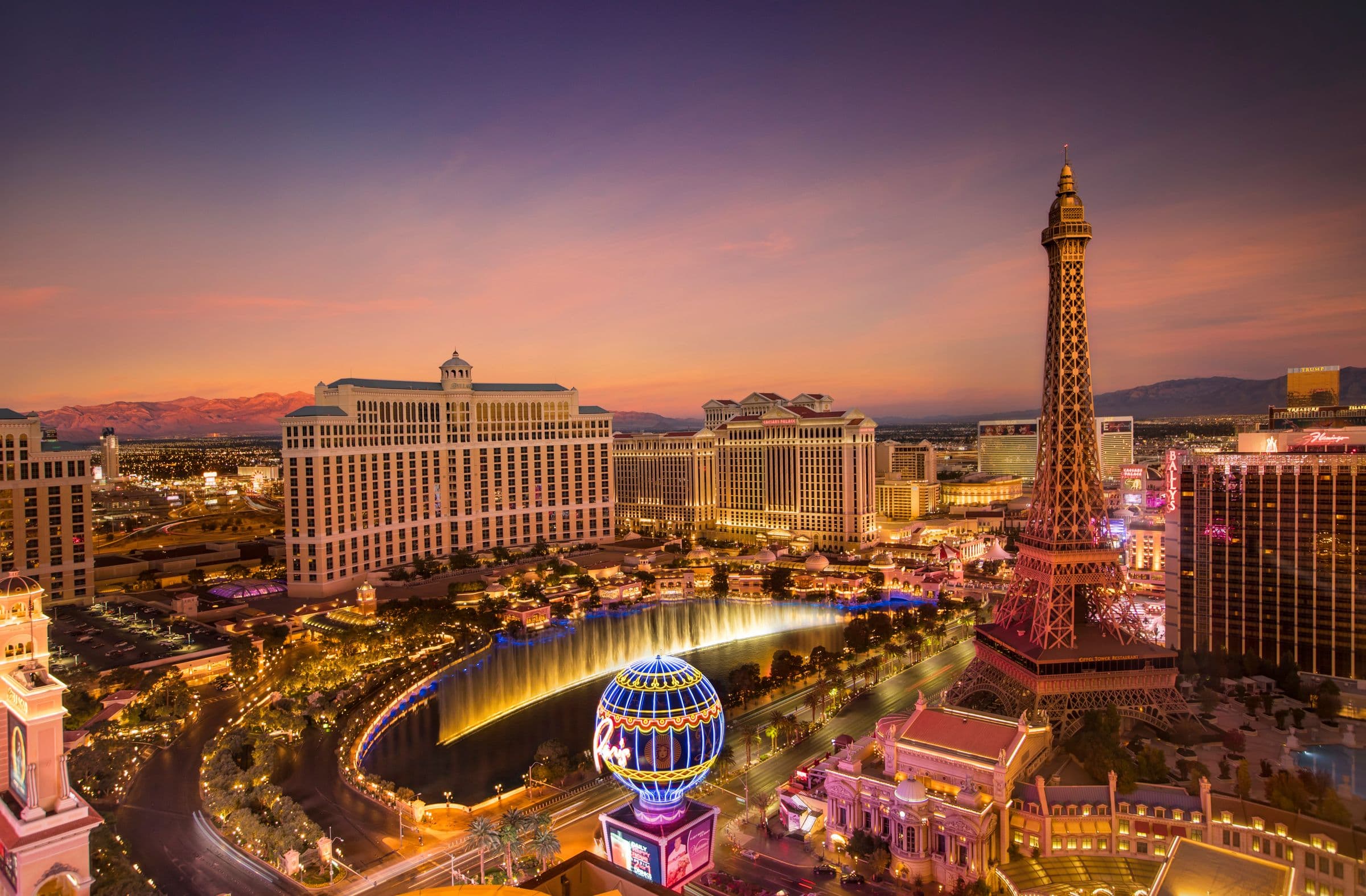 Aerial view of the Las Vegas Strip at dusk, with the Bellagio fountains and the Paris Eiffel Tower lit against a sunset sky