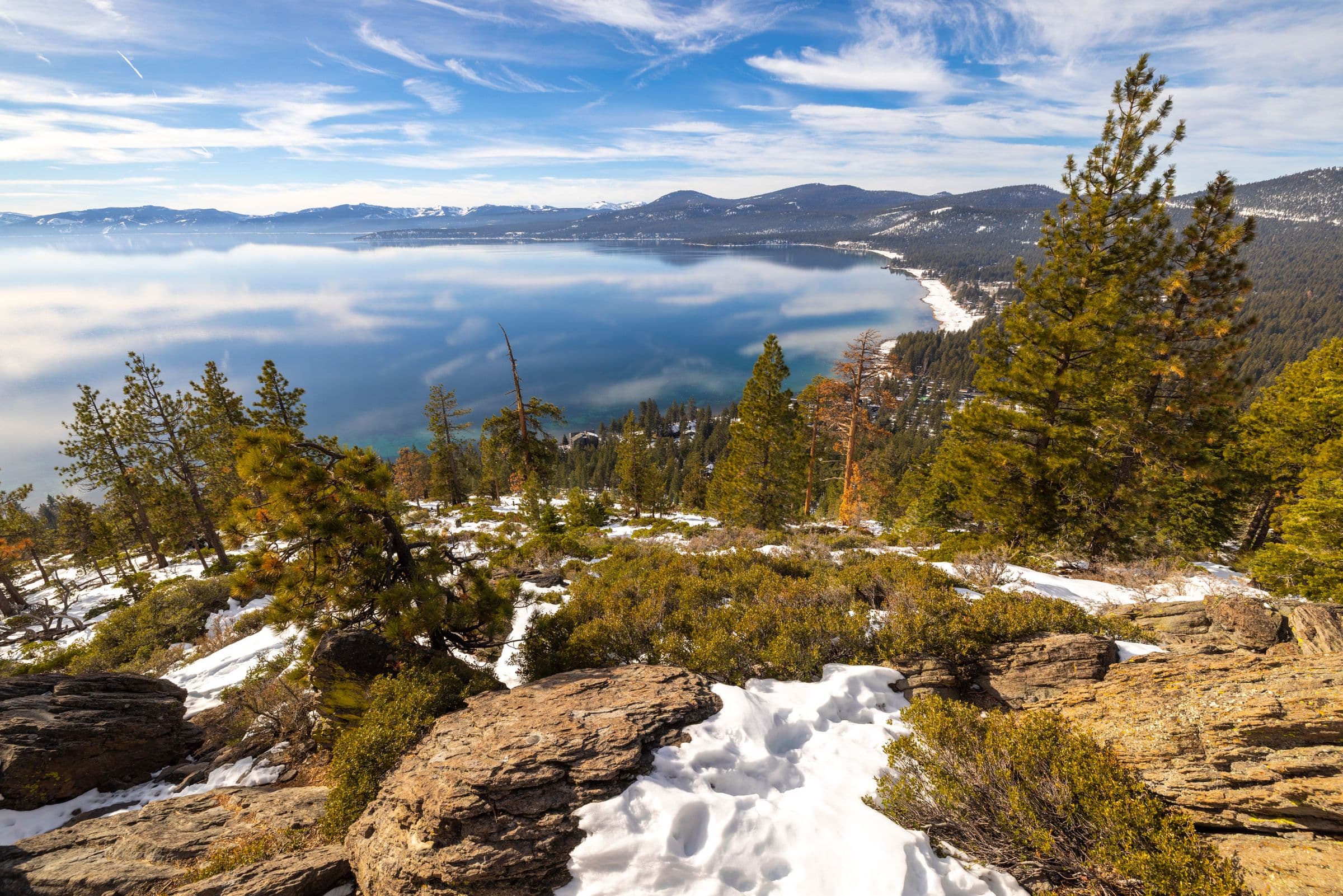 Lake Tahoe seen from a snowy Sierra Nevada overlook, with pine trees in the foreground and mountains across the water